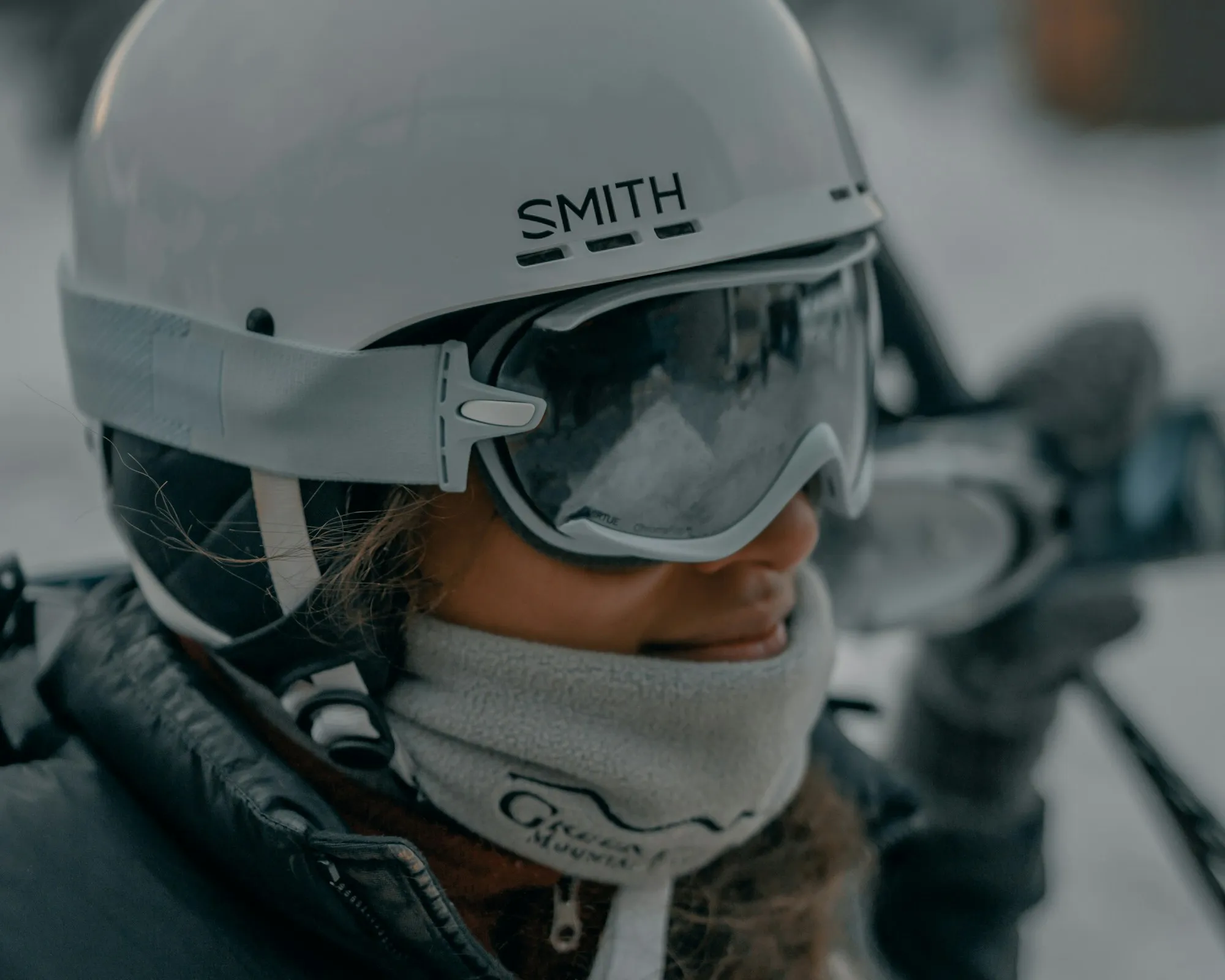 A skier in a white Smith helmet and mirrored Smith goggles with a fleece neck gaiter, ski poles behind, on an overcast day.