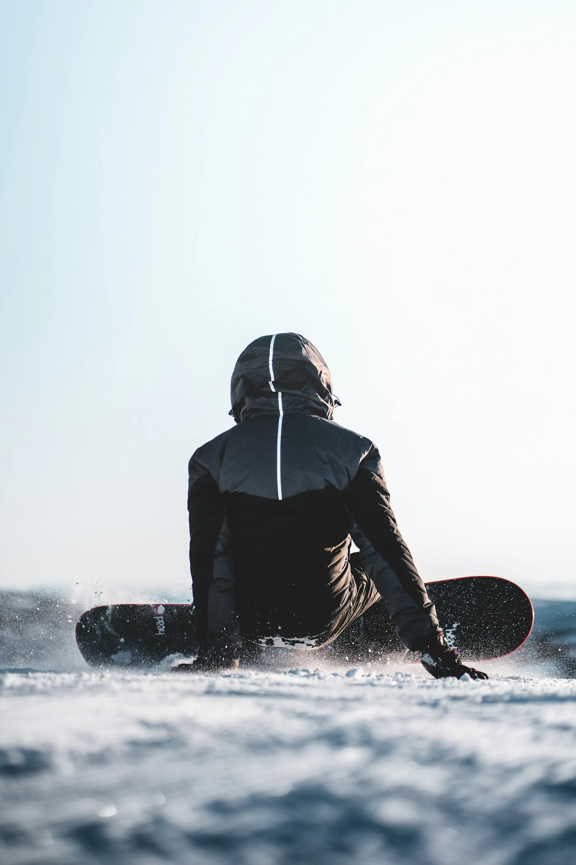 A snowboarder sitting back into a turn on a bright day, snow spraying off the edge, seen from behind against an open sky.