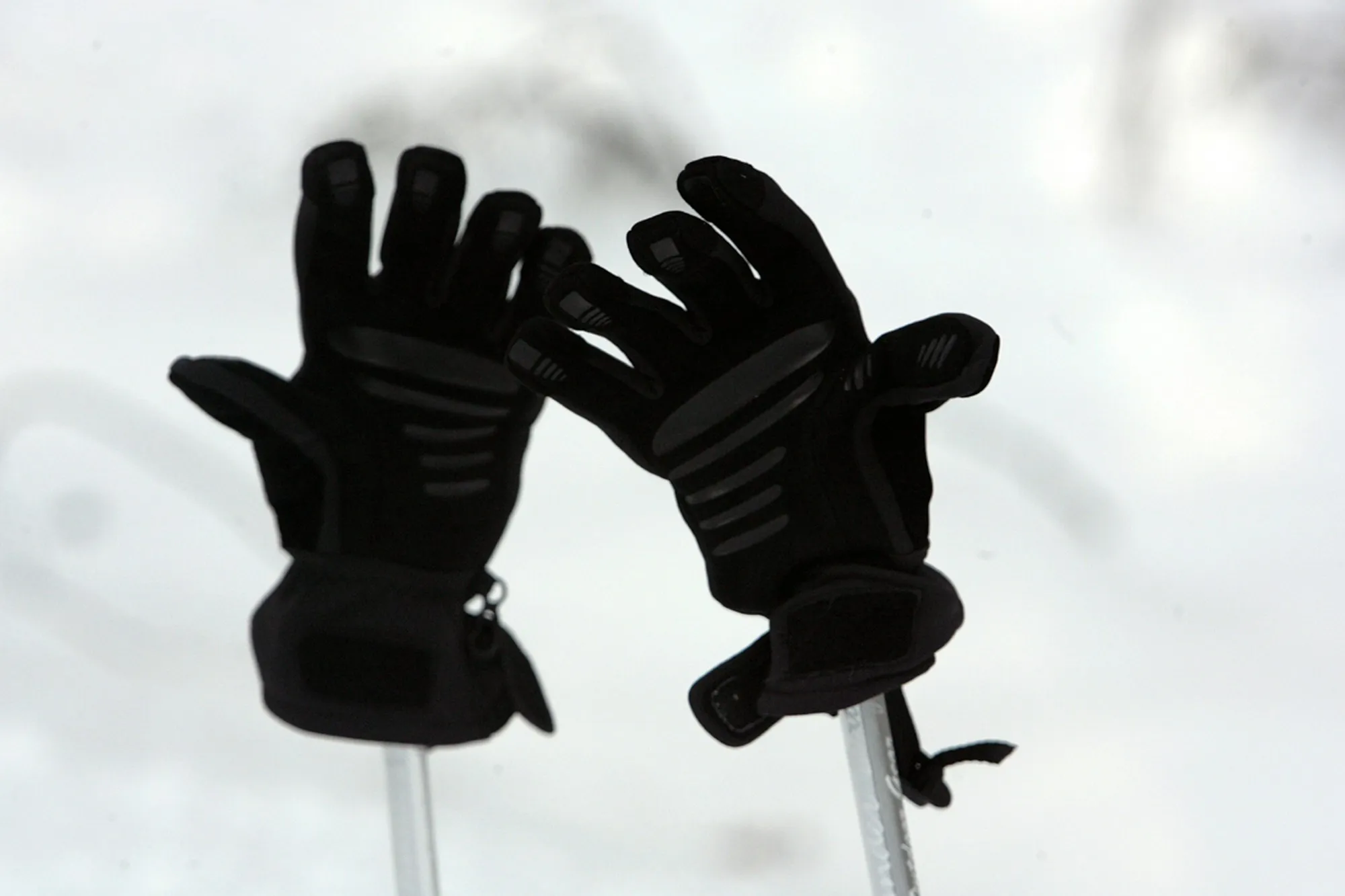 A pair of black insulated ski gloves resting fingers-up on ski poles against a snowy backdrop.