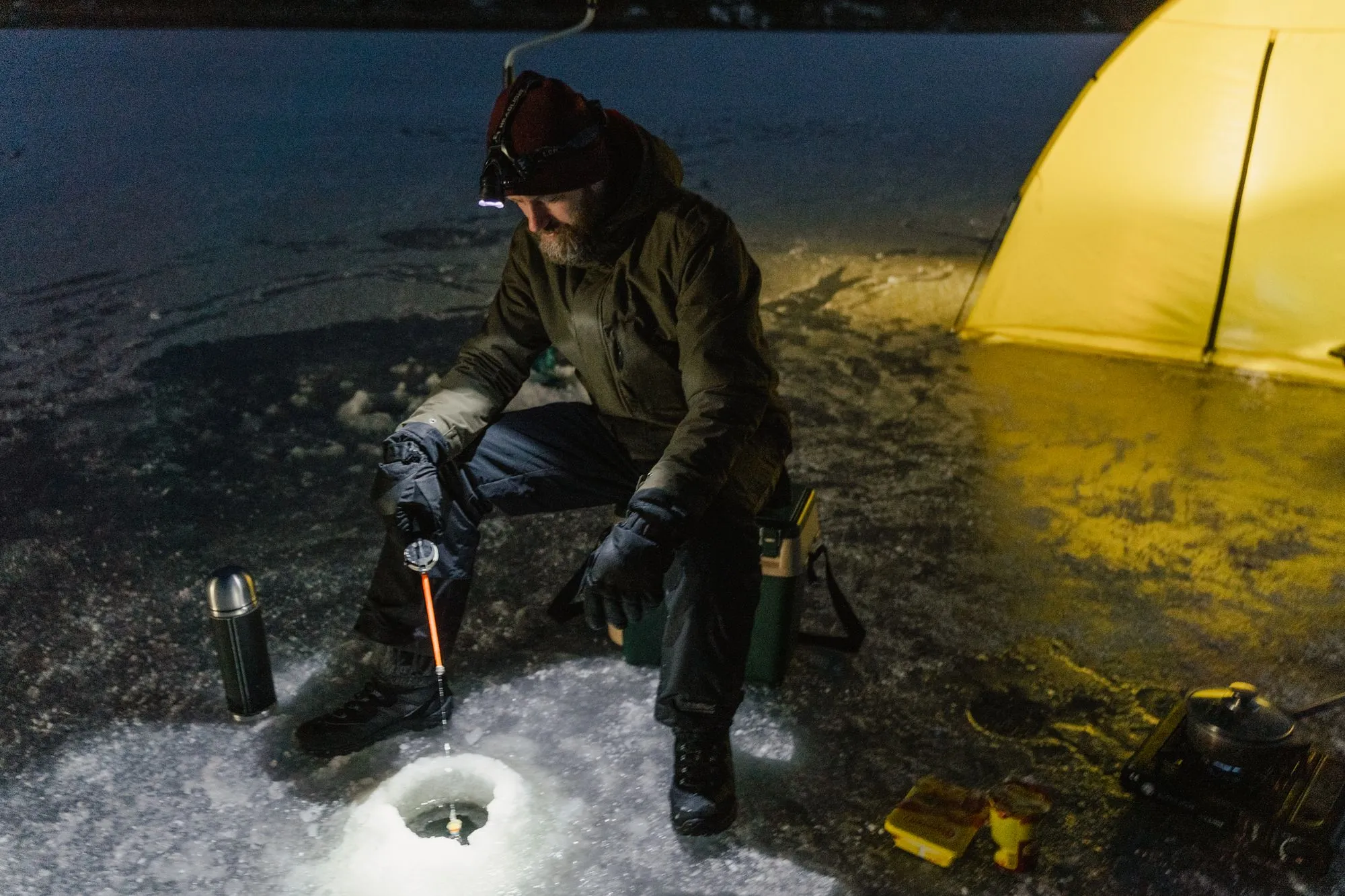 A man wearing a headlamp kneeling on a frozen lake at night, jigging a short rod over a drilled ice hole, a lit tent and a thermos beside him.